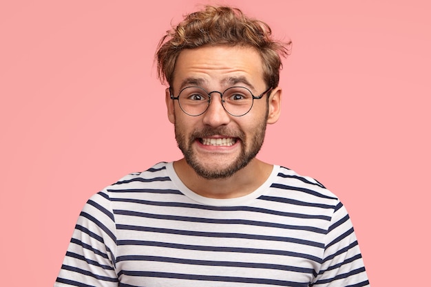 Free photo joyful caucasian male clenches teeth and looks positively, has curly hair, wears spectacles and striped sweater, isolated over pink wall. happy man freelancer rejoices success