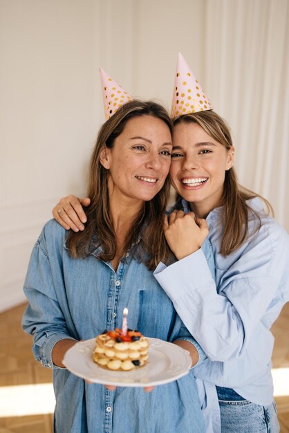 Joyful caucasian mother with her daughter wear shirts, stand in embrace and hold festive cake with candle. Holidays, celebration and women concept.