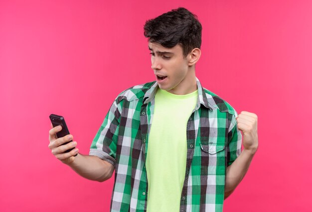 Радостный кавказский молодой человек в зеленой рубашке смотрит на телефон в руке и показывает жест да на изолированной розовой стене
