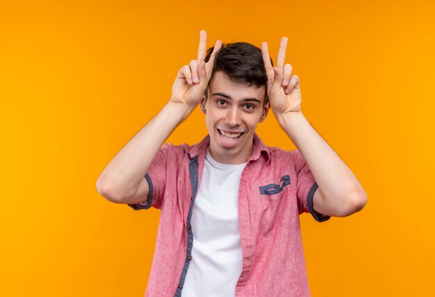 Joyful caucasian young man wearing pink shirt doing peace gesture with both hands and showing tongue on isolated orange wall