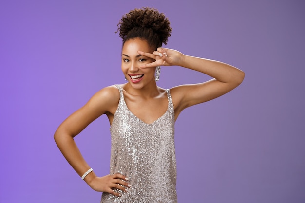 Joyful charming african-american woman in silver shiny evening dress smiling lively entertained grinning show victory peace gesture hold hand waist enjoying awesome party, blue background.