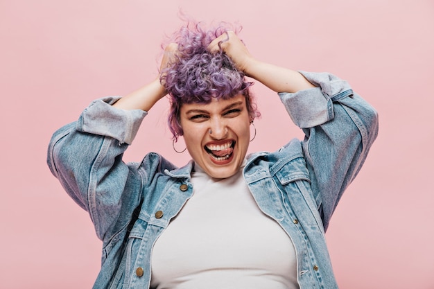 Joyful charming woman holds on to purple short hair in round earrings and in loose denim jacket on isolated pink.