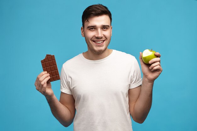 Радостный веселый молодой темноволосый парень смотрит в камеру с широкой взволнованной улыбкой и держит наполовину надкушенное зеленое яблоко