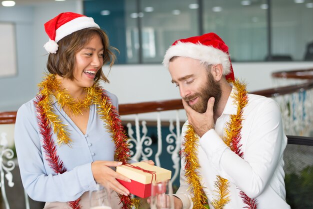 Joyful colleagues in Christmas hats exchanging gifts