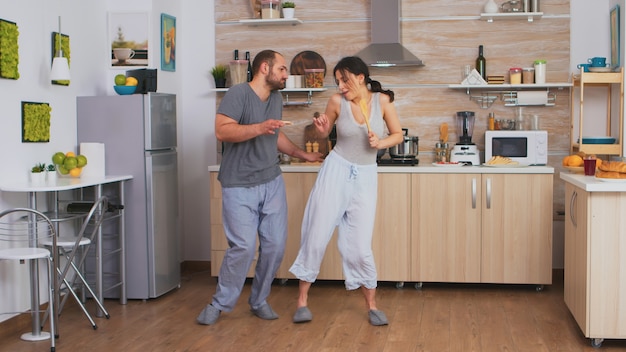 Joyful couple dancing and singing during breakfast in kitchen wearing pajamas. Carefree wife and husband laughing having fun funny enjoying life authentic married people positive happy relation