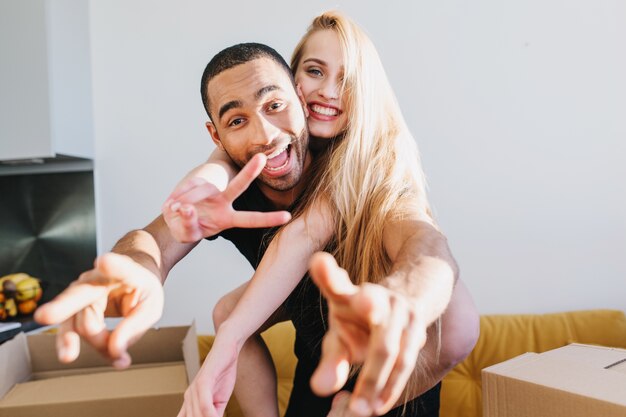 Joyful couple having fun in new house, just bought apartment, flat, unpacking boxes with their stuff. Young girl sitting on man's back, they showing peaces.