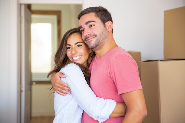 Joyful couple moving into new house, standing among carton boxes and hugging