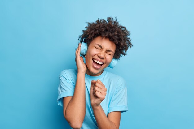 Joyful curly Afro American teenage girl holds hand near mouth like microphone sings favorite song loudly wears stereo headphones enjoys music wears casual t shirt isolated over blue wall