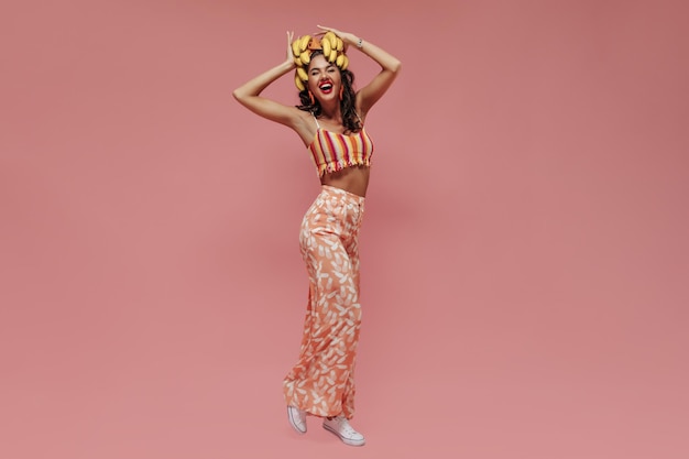 Joyful curly brunette haired girl witj cool earrings in modern top and wide pineapple print pants laughing and holding bananas