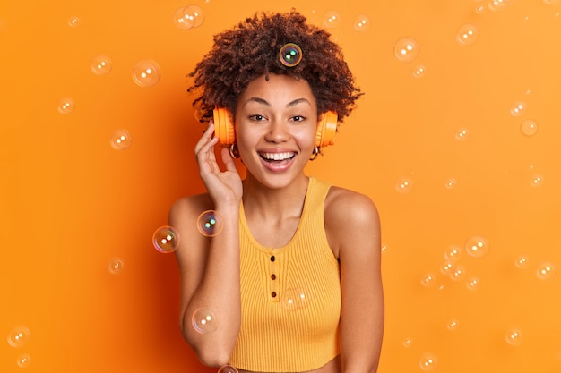 Joyful curly girl enjoys listening music via wireless headphones smiles gently enjoys spare time dressed in casual top isolated over orange wall soap bubbles flying around