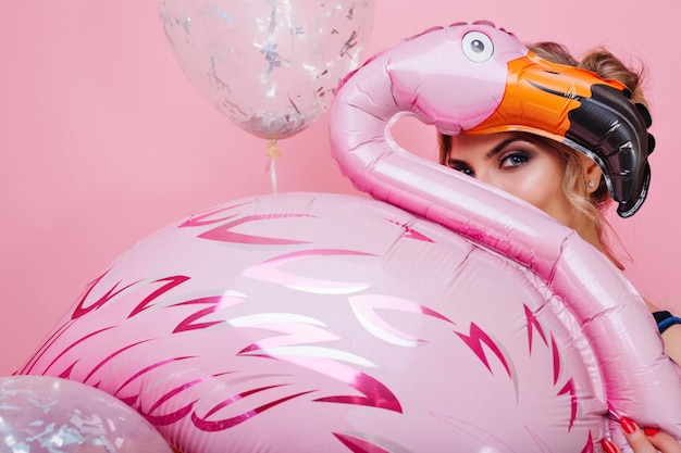 Joyful curly girl looking playfully hiding behind a big toy flamingo, standing on bright background. Funny young woman with trendy make-up posing holding glitter party balloon and pink inflatable bird