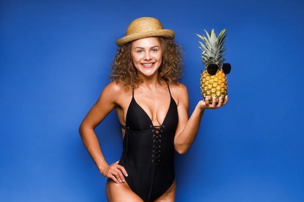 Joyful curly hair woman holding fresh pineapple with sunglasses, blue.