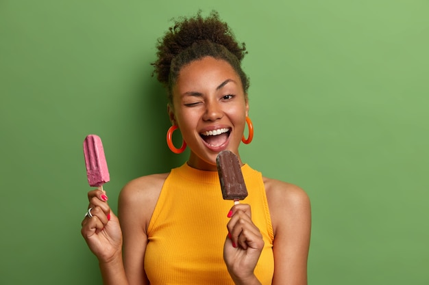 Free photo joyful curly haired woman bites delicious ice cream winks eye has fun happy expression
