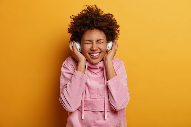 Joyful curly haired woman enjoys every bit of favorite song, listens music in stereo headphones, closes eyes and smiles broadly, inspired with awesome music, dressed in casual wear, stands indoor