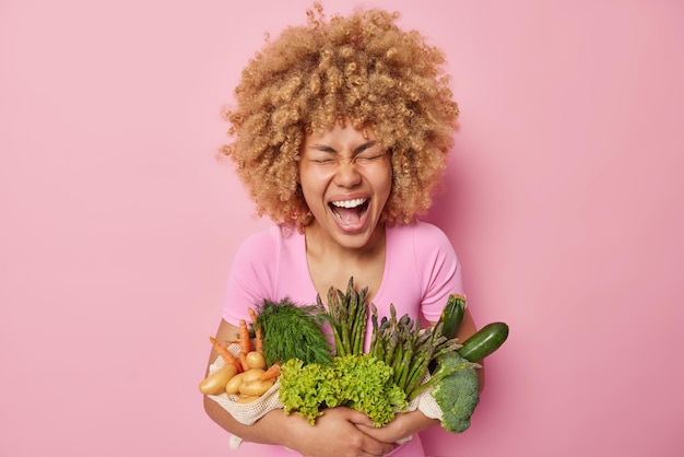 ビタミンでいっぱいの健康的な有機食品でポーズをとるうれしそうな縮れ毛の女性は、ベジタリアンサラダを調理するために行くピンクの背景の上に分離された幸福から生の新鮮な野菜を叫びます