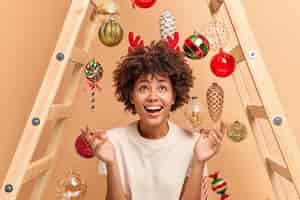 Free photo joyful curly haired woman raises hands looks above dressed casually wears reindeer red horns poses against beige background with new year toys hanging on ladder. holiday celebration concept.