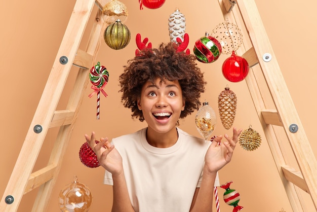 Joyful curly haired woman raises hands looks above dressed casually wears reindeer red horns poses against beige background with new year toys hanging on ladder. Holiday celebration concept.