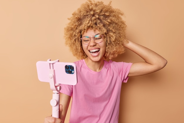 Free photo joyful curly haired woman uses selfie stick and smartphone for taking pictures laughs happily wears spectacles casual pink t shirt isolated over beige background joyful female photographs herself