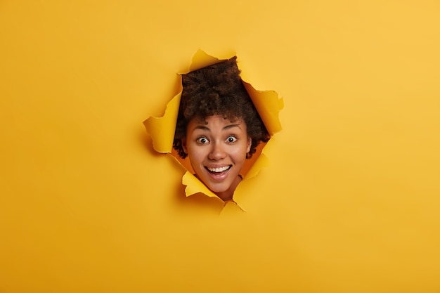 Joyful curly haired young woman keeps head in ripped paper hole, looks in wall breakthrough, smiles broadly, shows white perfect teeth