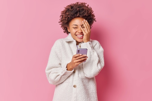 Joyful curly haired young woman makes face palm and smiles gladfully holds mobile phone reads funny message content wears warm white fur coat isolated over pink wall