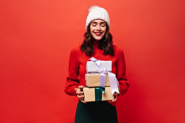 Joyful curly woman in bright red sweater, knitted hat poses closed eyes and holds gift boxes on isolated red wall