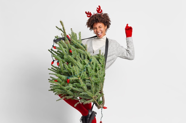 Joyful curly woman dances carefree has fun before New Year concert holds green fir tree as if guitar raises arm has playful mood happy to stay at home alone wears reindeer horns. Winter holidays