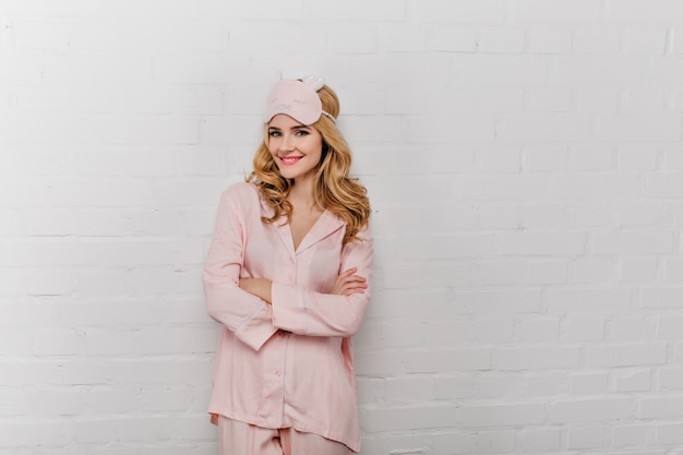Joyful curly woman in silk pajama standing in confident pose near bricked wall. Positive lady in eyemask smiling on white wall.