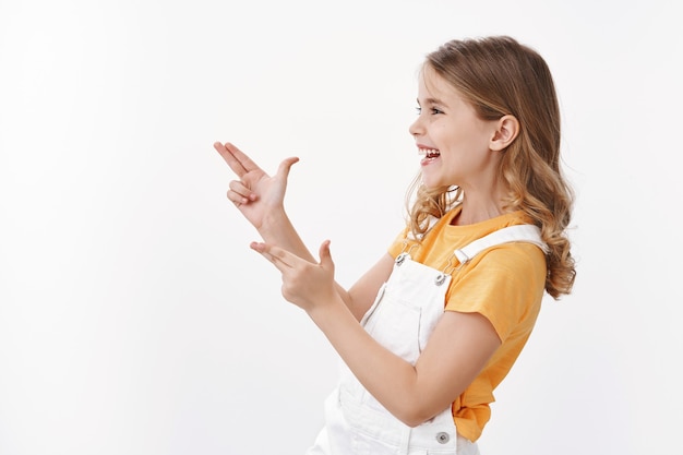 Joyful cute little girl having fun playing with friends, pointing finger pistols, looking make bang-bang sign left smiling and laughing playfully, enjoy summer holidays, standing white wall