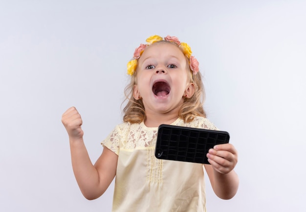 A joyful cute little girl wearing yellow shirt in floral headband raising clenched fist while holding mobile phone on a white wall