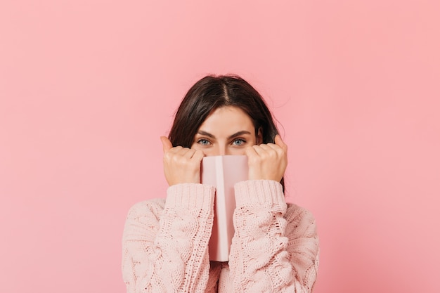 Foto gratuita gioiosa ragazza dai capelli scuri si copre il viso con il suo taccuino rosa. la donna in maglione lavorato a maglia esamina la macchina fotografica.