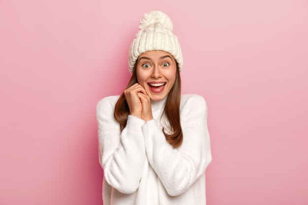 Joyful dark haired millennial girl has happy reaction on good news, smiles broadly, wears warm winter hat and comfortable white sweater, has enthusiastic gaze, isolated on pink wall