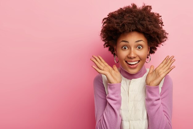 Joyful dark skinned female model raises hands in success gesture, keeps palms sideways, smiles broadly, gets awesome present from friend, dressed in casual wear, poses over rosy wall, empty space