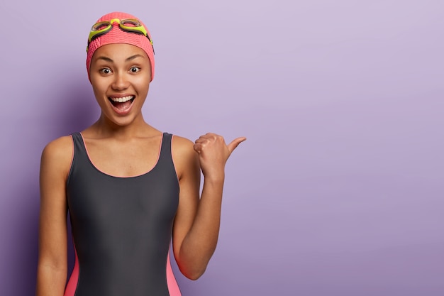 Joyful dark skinned female swimmer wears bathingcap, black swimsuit