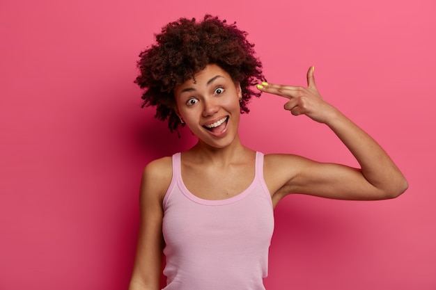 Joyful dark skinned girl shoots in temple, makes finger pistols, tilts head, has fun, wears casual vest, has positive expression, going crazy models against rosy wall bored during self isolation