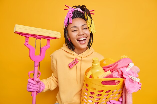 Joyful dark skinned housemaid smiles broadly wears sweatshirt and protective rubber gloves holds laundry basket and mop happy to finish housework isolated on yellow 