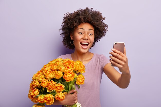 Joyful dark skinned lady expresses sincere feelings and emotions, makes selfie for sharing photo in social network, holds nice big bouquet of orange flowers, wears casual t shirt,