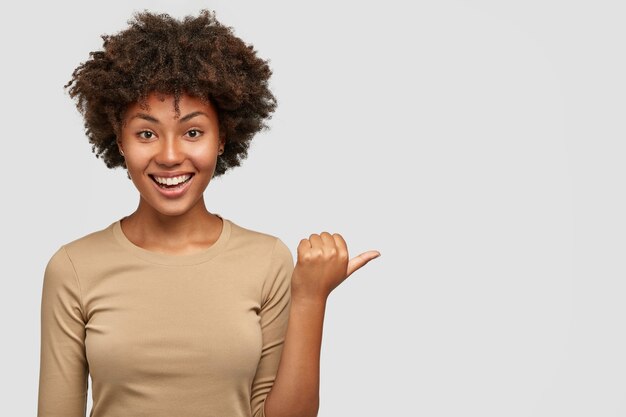 Joyful dark skinned smiling female has dark hair and curly hair, points aside with thumb