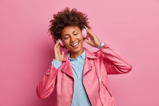 Joyful dark skinned woman has good mood listens music in stereo headphones closes eyes