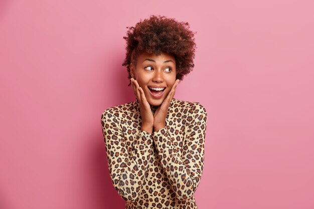 Joyful dark skinned woman keeps hands on cheeks, looks happily aside, has toothy smile, dressed in leopard turtleneck