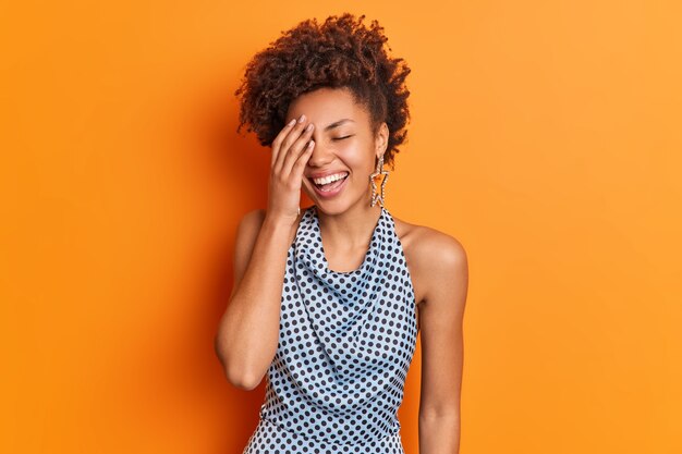 Joyful dark skinned woman makes face palm smiles pleasantly expresses positive emotions cannot stop laughing