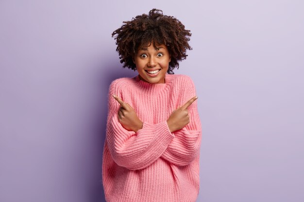 Joyful dark skinned woman with satisfied facial expression, curly hair, points sideways, keeps arms crossed over chest, being in high spirit, dressed in oversized rosy sweater, isolated on purple wall