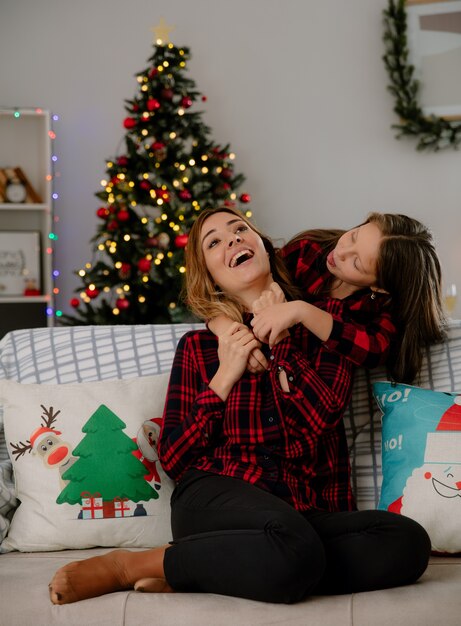 Joyful daughter stucks out tongue and hugs mother sitting on couch from behind enjoying christmas time at home