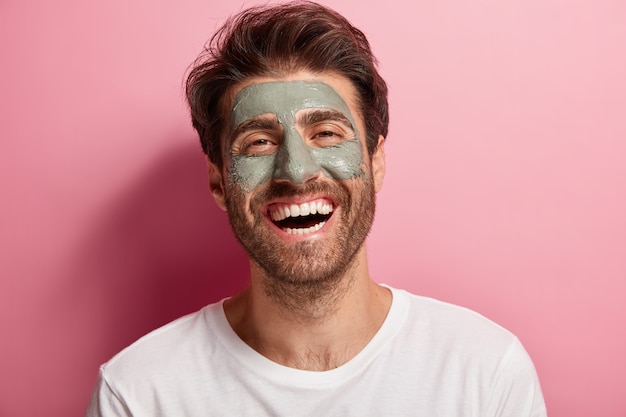 Joyful delighted man has clay mask on face, enjoys spa treatments, has broad smile, being in high spirit, cares about beauty