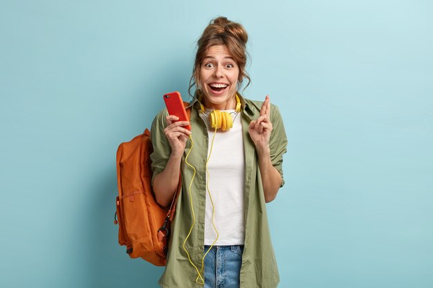 Радостная довольная женщина носит наушники, слушает музыку из приложения на современном мобильном телефоне, скрещивает пальцы на удачу