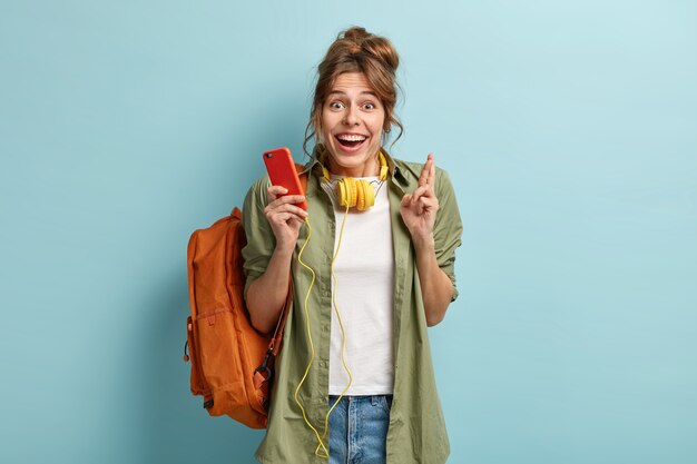 Joyful delighted woman wears headphones, listens music from app on modern cell phone, crosses fingers for good luck