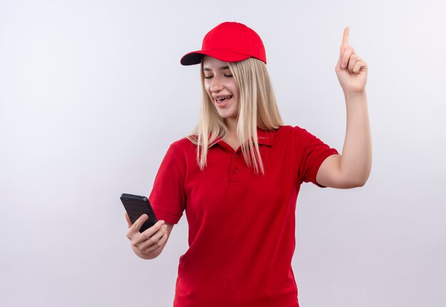 빨간 티셔츠와 모자를 쓰고 즐거운 배달 젊은 여자가 그녀의 손에 전화를보고 격리 된 흰 벽 에까지 가리 킵니다.