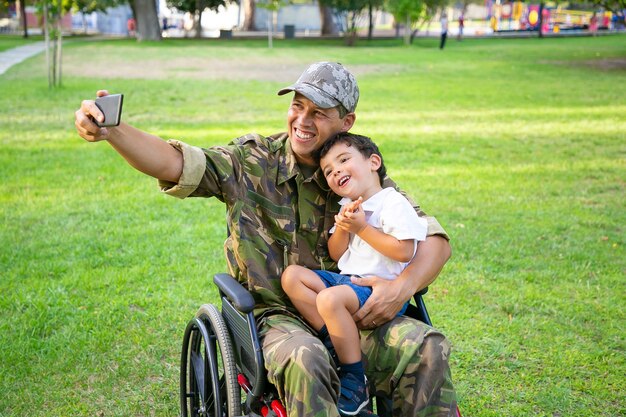 Радостный военный папа-инвалид и его маленький сын вместе делают селфи в парке. Мальчик сидит на коленях пап. Ветеран войны или концепция инвалидности