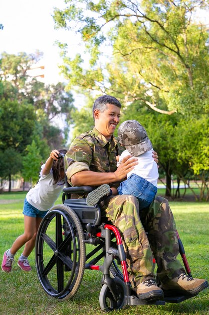 Joyful disabled military dad walking with two children in park. Girl pushing wheelchair handles, boy sitting on dads lap. Veteran of war or disability concept