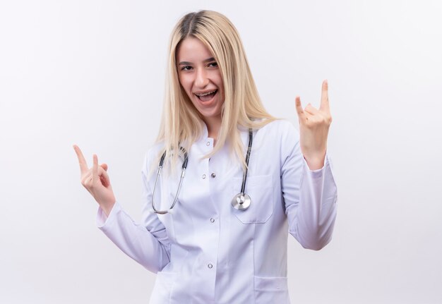 Радостный доктор молодая блондинка носить стетоскоп и медицинский халат в стоматологической скобе, показывая козлиный жест обеими руками на изолированной белой стене