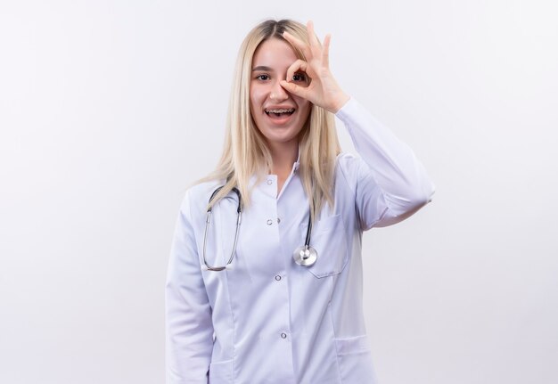 Joyful doctor young blonde girl wearing stethoscope and medical gown in dental brace showing look gesture on isolated white wall